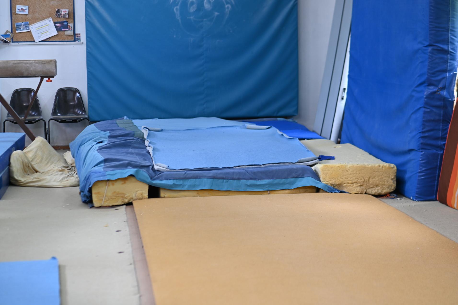 Kleine Turnarena mit gestapelten Matten, blauer Polsterung und Stühlen im Hintergrund.