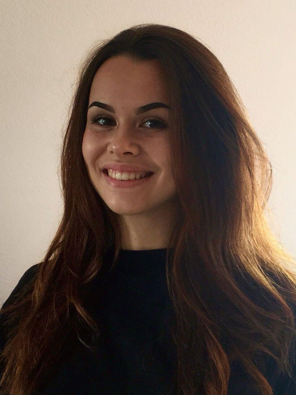 Portrait einer lächelnden Frau mit langen, braunen Haaren, vor einem hellen Hintergrund.