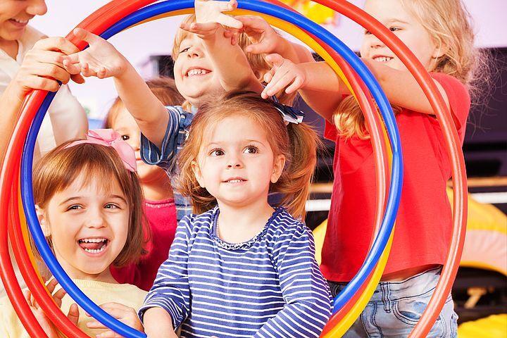 Sechs Kinder stehen lächelnd in einem bunten Ringspiel, während ein Mädchen in der Mitte den Fokus bildet.